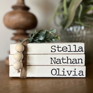May include: A stack of three white wooden blocks with the names Stella, Nathan, and Olivia written in black lettering. The blocks are decorated with a brown twine bow and a string of wooden beads.