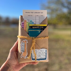 Peut inclure: Un colis en papier brun envelopp&eacute; de ficelle et d&eacute;cor&eacute; d'autocollants. Les autocollants repr&eacute;sentent une girafe portant une pile de livres, un livre avec le texte "The Book Was Better" et une carte de biblioth&egrave;que avec le texte "From the Library Of".
