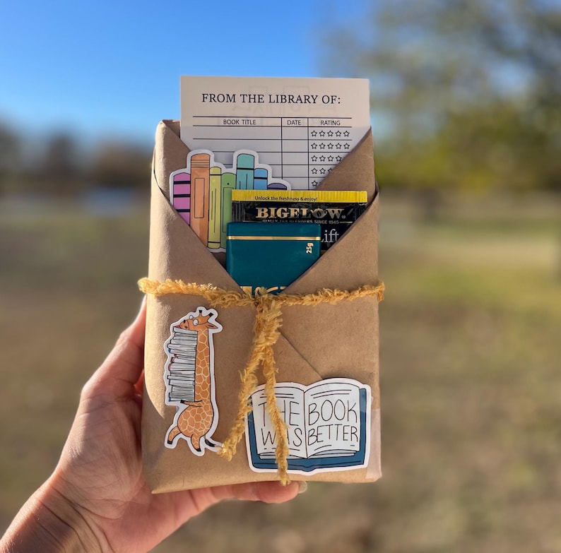 Peut inclure: Un colis en papier brun envelopp&eacute; de ficelle et d&eacute;cor&eacute; d'autocollants. Les autocollants repr&eacute;sentent une girafe portant une pile de livres, un livre avec le texte "The Book Was Better" et une carte de biblioth&egrave;que avec le texte "From the Library Of".