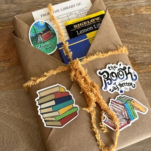 Peut inclure: Un colis en papier kraft ficel&eacute; avec une ficelle jaune. Le colis contient un livre, des sachets de th&eacute; et des autocollants. Les autocollants repr&eacute;sentent une pile de livres, un livre avec le texte "The Book Was Better" et une sc&egrave;ne de camping avec le texte "Adventure awaits". La bo&icirc;te de sachets de th&eacute; indique "Bigelow Lemon Lift".