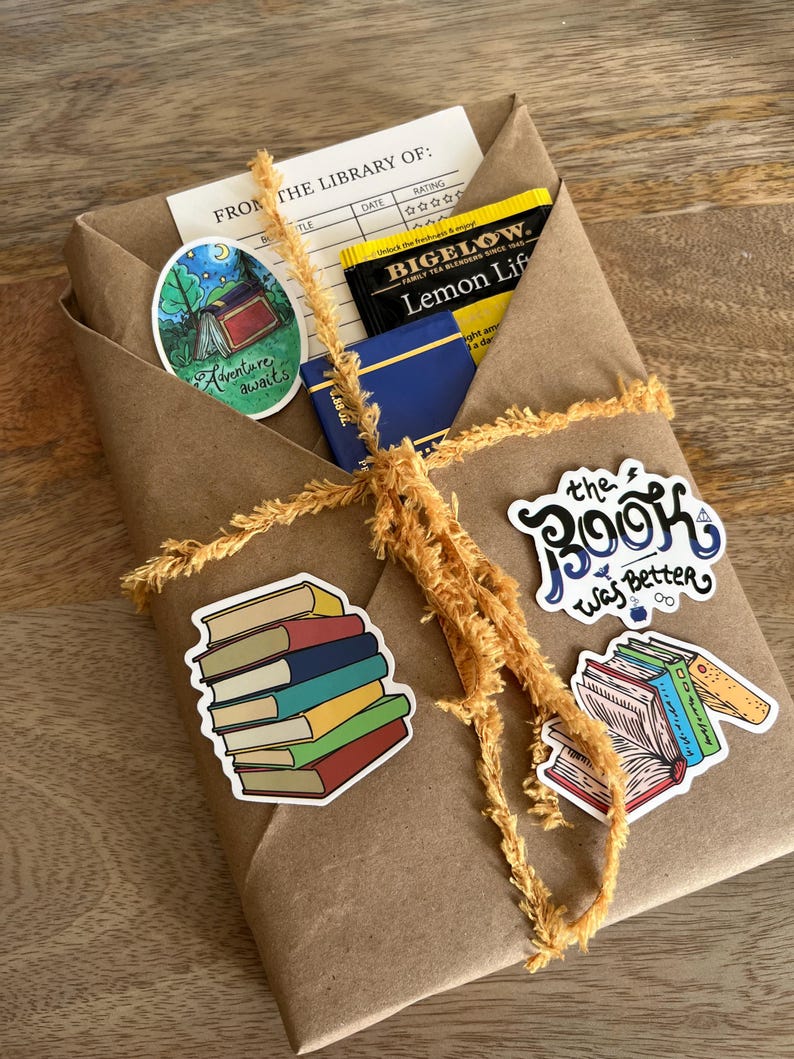 Peut inclure: Un colis en papier kraft ficel&eacute; avec une ficelle jaune. Le colis contient un livre, des sachets de th&eacute; et des autocollants. Les autocollants repr&eacute;sentent une pile de livres, un livre avec le texte "The Book Was Better" et une sc&egrave;ne de camping avec le texte "Adventure awaits". La bo&icirc;te de sachets de th&eacute; indique "Bigelow Lemon Lift".