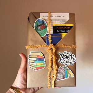 Peut inclure: Un colis en papier kraft envelopp&eacute; de ficelle et d&eacute;cor&eacute; d'autocollants color&eacute;s. Les autocollants repr&eacute;sentent une pile de livres, une sc&egrave;ne de camping avec le texte "Adventure awaits", une bo&icirc;te de sachets de th&eacute; avec le texte "BIGELOW. Lemon Lift", et un livre avec le texte "the Book was Better".