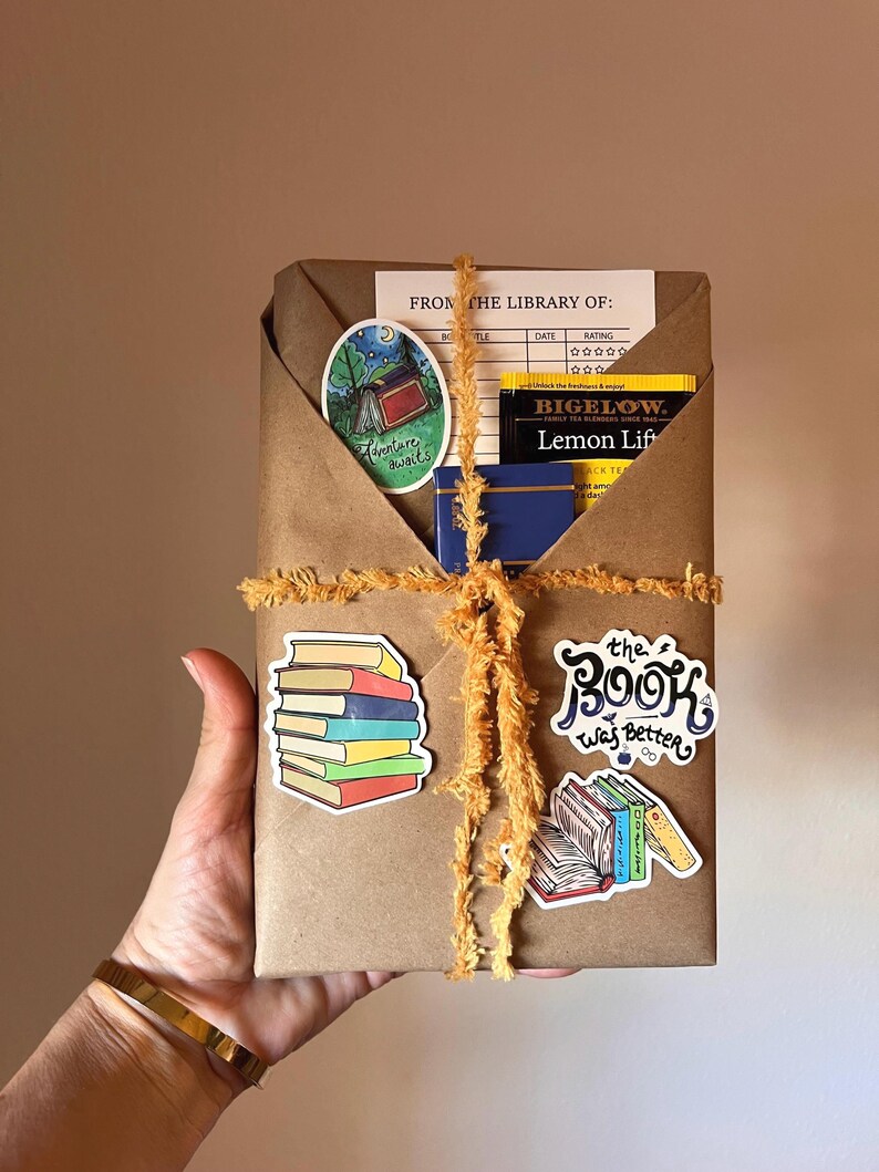 Peut inclure: Un colis en papier kraft envelopp&eacute; de ficelle et d&eacute;cor&eacute; d'autocollants color&eacute;s. Les autocollants repr&eacute;sentent une pile de livres, une sc&egrave;ne de camping avec le texte "Adventure awaits", une bo&icirc;te de sachets de th&eacute; avec le texte "BIGELOW. Lemon Lift", et un livre avec le texte "the Book was Better".