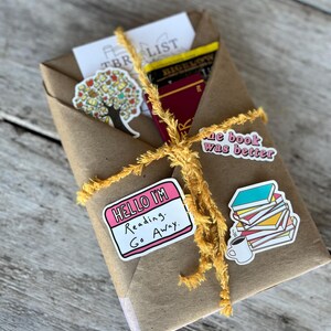 Peut inclure: Un colis en papier brun envelopp&eacute; de ficelle jaune et d&eacute;cor&eacute; d'autocollants. Les autocollants comprennent un arbre avec des feuilles, un autocollant rose qui dit "Hello I'm Reading. Go Away." et un autocollant d'une pile de livres avec une tasse de caf&eacute;.