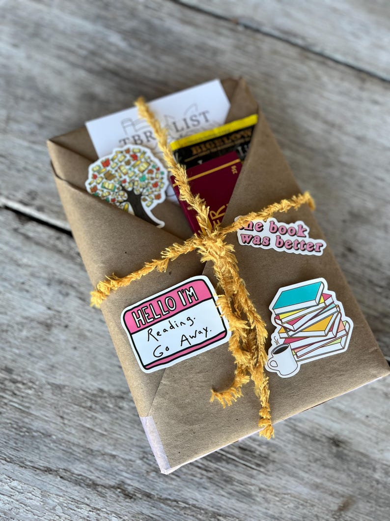 Peut inclure: Un colis en papier brun envelopp&eacute; de ficelle jaune et d&eacute;cor&eacute; d'autocollants. Les autocollants comprennent un arbre avec des feuilles, un autocollant rose qui dit "Hello I'm Reading. Go Away." et un autocollant d'une pile de livres avec une tasse de caf&eacute;.