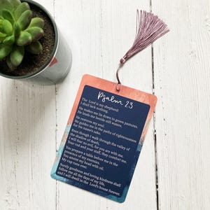 Pode incluir: Um marcador retangular com o texto do Salmo 23, com fundo azul marinho e borda rosa e laranja. Uma borla malva está presa no topo. No fundo, há uma pequena planta suculenta em vaso.