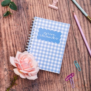 May include: A spiral-bound notebook with a blue and white gingham pattern. The cover features a blue cloud-shaped label with the words "give thanks" in script. A pink rose, pencils, and paper clips are arranged around the notebook on a wooden surface.
