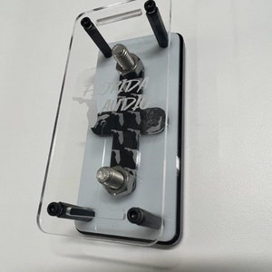 May include: A clear acrylic phone stand with black metal accents. The stand has a white background with the text "Florida Audio" printed on it.