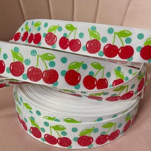 May include: A white grosgrain ribbon featuring a repeating pattern of bright red cherries with green stems and leaves, interspersed with small teal polka dots. The ribbon has a textured, ribbed surface and is wound into a tight, circular spool.