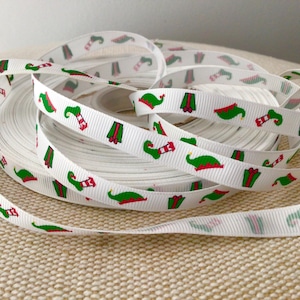 May include: White ribbon with a repeating pattern of green elf hats, red and green gift boxes, and red and green striped stockings. The ribbon is rolled up in a coil. The ribbon is likely used for gift wrapping or crafting.