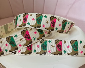 Op de afbeelding: Een wit grosgrain lint met een herhalend patroon van bruine cowboylaarzen met glitterende roze of turquoise accenten, afgewisseld met kleine gouden en roze sterren. Het lint heeft een getextureerd oppervlak en is in meerdere lagen opgerold.
