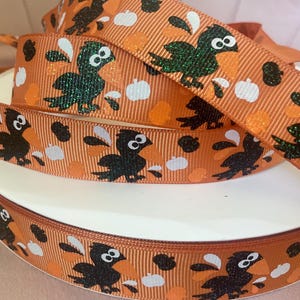 May include: Orange ribbon with a repeating pattern of black crows, white pumpkins, and orange dots. The crows have glittery wings. The ribbon is wrapped around a white spool, perfect for crafting and seasonal decorations.