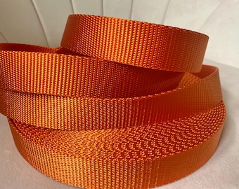 May include: A roll of vibrant orange nylon webbing featuring a tight, consistent woven texture. The material is coiled into a circular shape, displaying a smooth, durable surface with a distinct cross-hatched pattern throughout the entire length.
