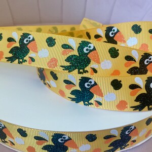 May include: Yellow ribbon with a repeating pattern of cartoon crows with orange beaks and accents. The crows are in various shades of green and black, with white and orange shapes scattered throughout the design. The ribbon is curled.