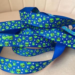 Op de afbeelding: Blauw lint met een herhaald patroon van groene alien gezichten en handen. Het lint heeft effen blauwe randen en een getextureerd oppervlak. Geschikt voor knutselen en cadeauverpakking. Het lint is ongeveer 2,5 cm breed.