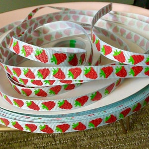 May include: A roll of white ribbon with a repeating pattern of red strawberries with green leaves. The ribbon is on a white surface.