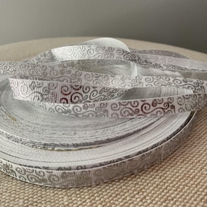 May include: A roll of white ribbon with a silver, swirling pattern. The ribbon is partially unrolled, revealing the intricate design. The ribbon is likely used for crafts, gift wrapping, or decorative purposes.