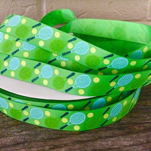 May include: A spool of bright green ribbon with a repeating pattern of tennis rackets and yellow tennis balls. The tennis rackets are light blue with black handles. The ribbon is wrapped around a white cardboard spool.