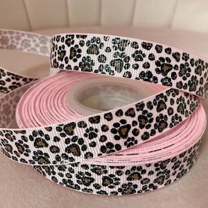 May include: Pink ribbon spooled on a clear plastic core, with a white background and a repeating pattern of black and brown paw prints. A length of the ribbon is unspooled, showing the paw print design.