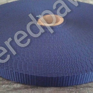 May include: A roll of dark blue webbing, a type of heavy-duty fabric tape, is wound around a cardboard core. The webbing is textured and has a woven appearance.