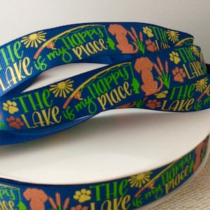 May include: Blue ribbon with the text "The Lake is my Happy Place." The ribbon features illustrations of a dog, paw prints, fishing poles, and suns. The ribbon is rolled on a white spool.