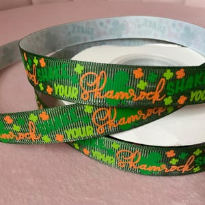 May include: Green ribbon with the words "Shake Your Shamrock" in orange script, accented with green and orange shamrocks. The ribbon is coiled on a white spool, perfect for St. Patrick's Day crafts and decorations.