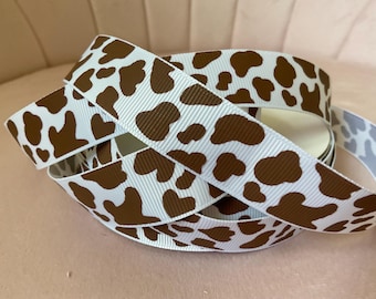 May include: A spool of white grosgrain ribbon featuring a repeating brown cow print pattern. The ribbon is coiled in multiple loops, displaying the textured fabric surface and the irregular brown spots against the solid white background throughout.