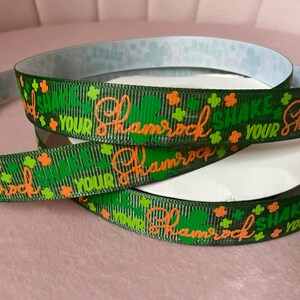 May include: Green ribbon with the text "Shake Your Shamrock" in orange script, along with green shamrock and orange clover designs. The ribbon is coiled, showing the printed design on the front and a patterned design on the back.
