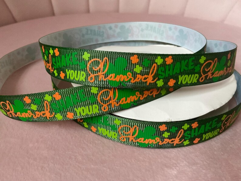 May include: Green ribbon with the text "Shake Your Shamrock" in orange script, along with green shamrock and orange clover designs. The ribbon is coiled, showing the printed design on the front and a patterned design on the back.