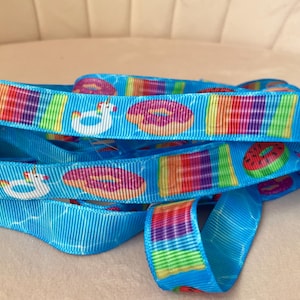 May include: A bright blue grosgrain ribbon featuring a repeating pattern of colorful pool floats, including pink frosted donuts, rainbow-striped rectangular rafts, watermelon slices, and white unicorn-shaped inflatable rings on a water-like background.