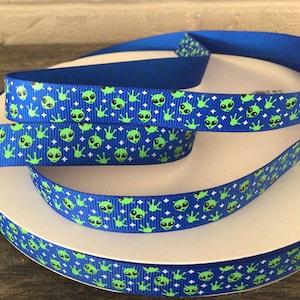 Op de afbeelding: Blauw lint met een herhalend patroon van groene aliengezichten en -handen op een blauwe achtergrond, afgewisseld met witte ruitvormen. Het lint is opgerold op een wit oppervlak.