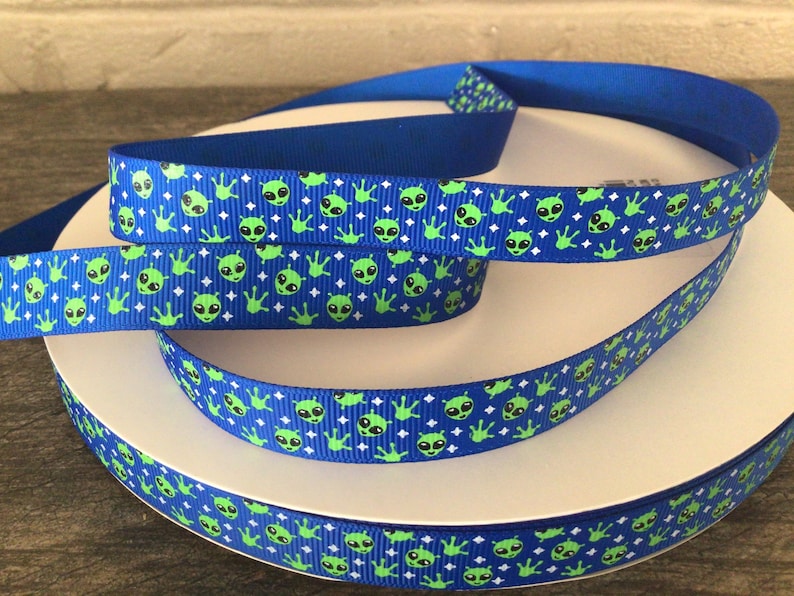 Op de afbeelding: Blauw lint met een herhalend patroon van groene aliengezichten en -handen op een blauwe achtergrond, afgewisseld met witte ruitvormen. Het lint is opgerold op een wit oppervlak.