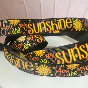 May include: A black grosgrain ribbon featuring a repeating pattern of yellow and orange sun illustrations and the text "You are my sunshine" printed in a stylized orange font. The ribbon is coiled, showing the continuous design on the fabric surface.