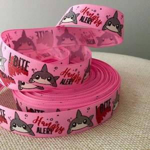 May include: Pink ribbon with a repeating pattern of cartoon sharks, the words "I BITE" in red, and "Hungry Alert" in red. The ribbon is rolled up and ready for use.