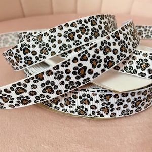 May include: White ribbon with a repeating pattern of brown and black paw prints. The ribbon is rolled and layered, showcasing the design. The ribbon has a slight sheen, adding a touch of elegance.