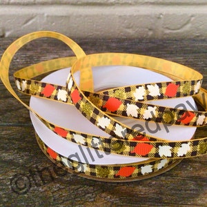 May include: A spool of decorative ribbon with a fall-themed pattern. The ribbon features a yellow background with a plaid design in brown and gold. The ribbon is adorned with orange, white, and gold leaf shapes. The ribbon is perfect for crafting and gift wrapping.