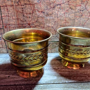 Könnte beinhalten: Zwei Messing-Pflanzgefäße mit glänzendem, goldenem Finish. Jedes Pflanzgefäß hat ein dekoratives Band mit blattartigen Designs in der Mitte. Die Pflanzgefäße stehen auf einer Holzoberfläche mit einer Karte im Hintergrund.
