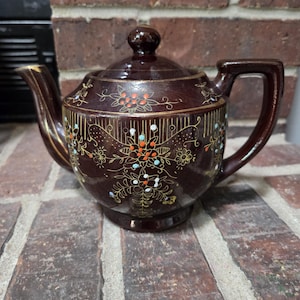 May include: A dark brown ceramic teapot with a matching lid, featuring a gold-lined floral pattern with small orange and light blue accents. The teapot has a curved spout, a sturdy handle, and a rounded body with a pedestal base.
