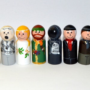 May include: A set of eight hand-painted wooden peg dolls depicting characters from the classic novel 'A Christmas Carol' by Charles Dickens. The dolls are painted in various colors, including white, green, red, black, and gray. The dolls are all wearing different costumes and accessories, including a top hat, a scarf, a chain, and a wreath. The dolls are arranged in a row on a white background.