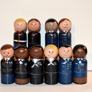 May include: A set of ten hand-painted wooden peg dolls dressed in military uniforms. The dolls are all wearing different uniforms, representing the different branches of the U.S. military. The dolls are all standing in a row, with the tallest doll in the center.