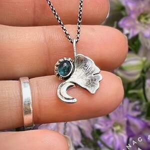 K&ouml;nnte beinhalten: Eine silberne Halskette mit einem Ginkgoblatt-Anh&auml;nger und einem kleinen blauen Edelstein. Der Anh&auml;nger hat eine Halbmondform und befindet sich an einer zarten Kette. Der Hintergrund zeigt lila Blumen.