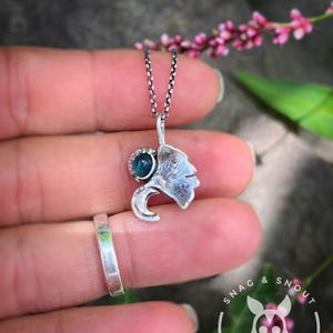 K&ouml;nnte beinhalten: Eine silberne Anh&auml;ngerkette mit einem Blattmotiv und einem kleinen, teal-farbenen Edelstein. Der Anh&auml;nger h&auml;ngt an einer feinen Kette. Ein silberner Ring ist ebenfalls zu sehen, zusammen mit einem Cartoon-Tier-Logo.