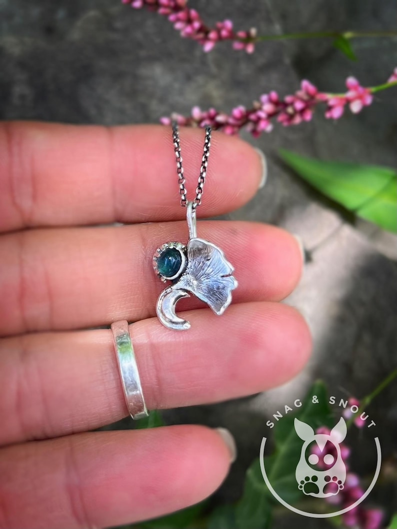 K&ouml;nnte beinhalten: Eine silberne Anh&auml;ngerkette mit einem Blattmotiv und einem kleinen, teal-farbenen Edelstein. Der Anh&auml;nger h&auml;ngt an einer feinen Kette. Ein silberner Ring ist ebenfalls zu sehen, zusammen mit einem Cartoon-Tier-Logo.