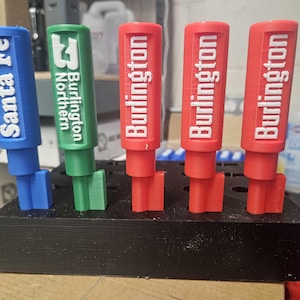 May include: A collection of colorful, 3D-printed train-themed items. The items include blue and white "Santa Fe" and green "Burlington Northern" pieces, along with several red "Burlington" pieces. They are displayed in a black holder.