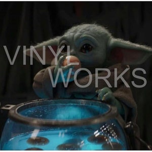 May include: A small, green, and grey alien-like creature with large ears is holding a small, round, orange object in its hand. The creature is sitting in front of a blue, glass container with a black rim. The container is filled with a blue liquid and several small, round, black objects.