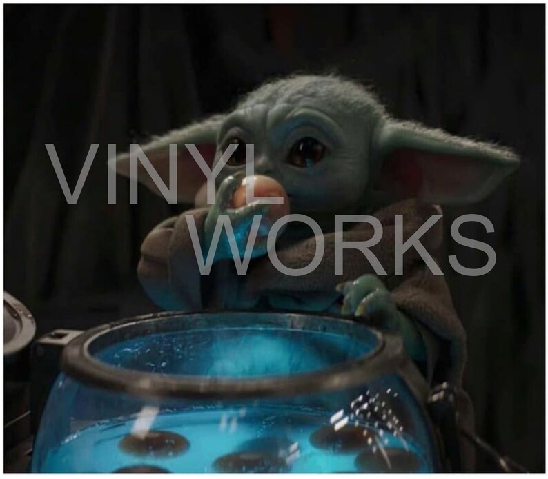 May include: A small, green, and grey alien-like creature with large ears is holding a small, round, orange object in its hand. The creature is sitting in front of a blue, glass container with a black rim. The container is filled with a blue liquid and several small, round, black objects.