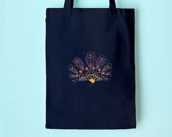 May include: A navy blue tote bag with a decorative design featuring a mountain range, sun, and stars in shades of orange and pink. The bag is hanging on a wooden peg against a light blue wall.