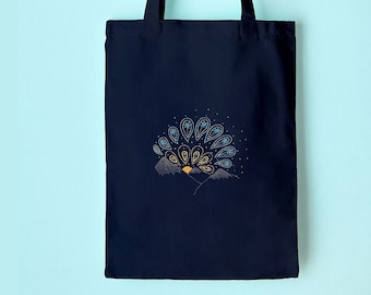 May include: A navy blue tote bag with a decorative design of a mountain range and a stylized sunburst in shades of gold, blue, and white. The bag is hanging on a wooden peg against a light blue wall.