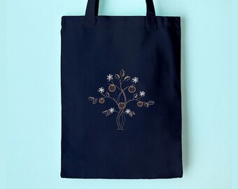 May include: A navy blue tote bag with a decorative embroidered design of a tree with fruit and star-shaped elements. The tote bag is hanging on a wooden peg against a light blue wall. The bag has two long handles.