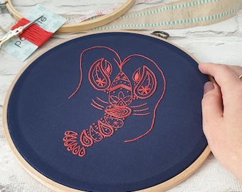 May include: A red embroidered lobster design on a dark blue fabric stretched over a wooden embroidery hoop.